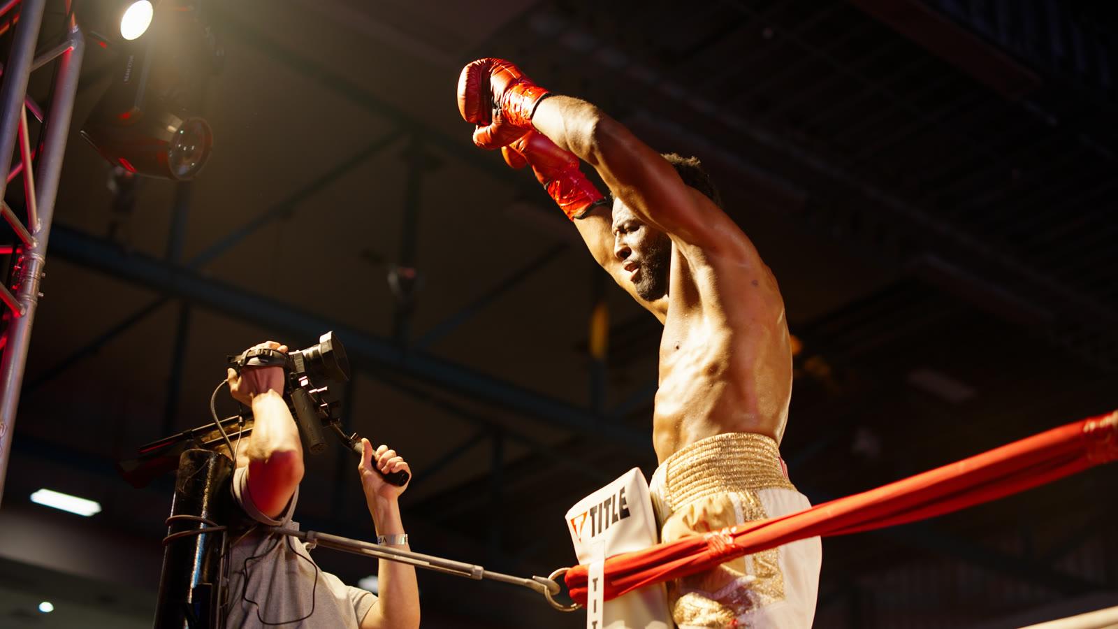 A victorious boxer in the ring, arms raised, as a camera operator films him