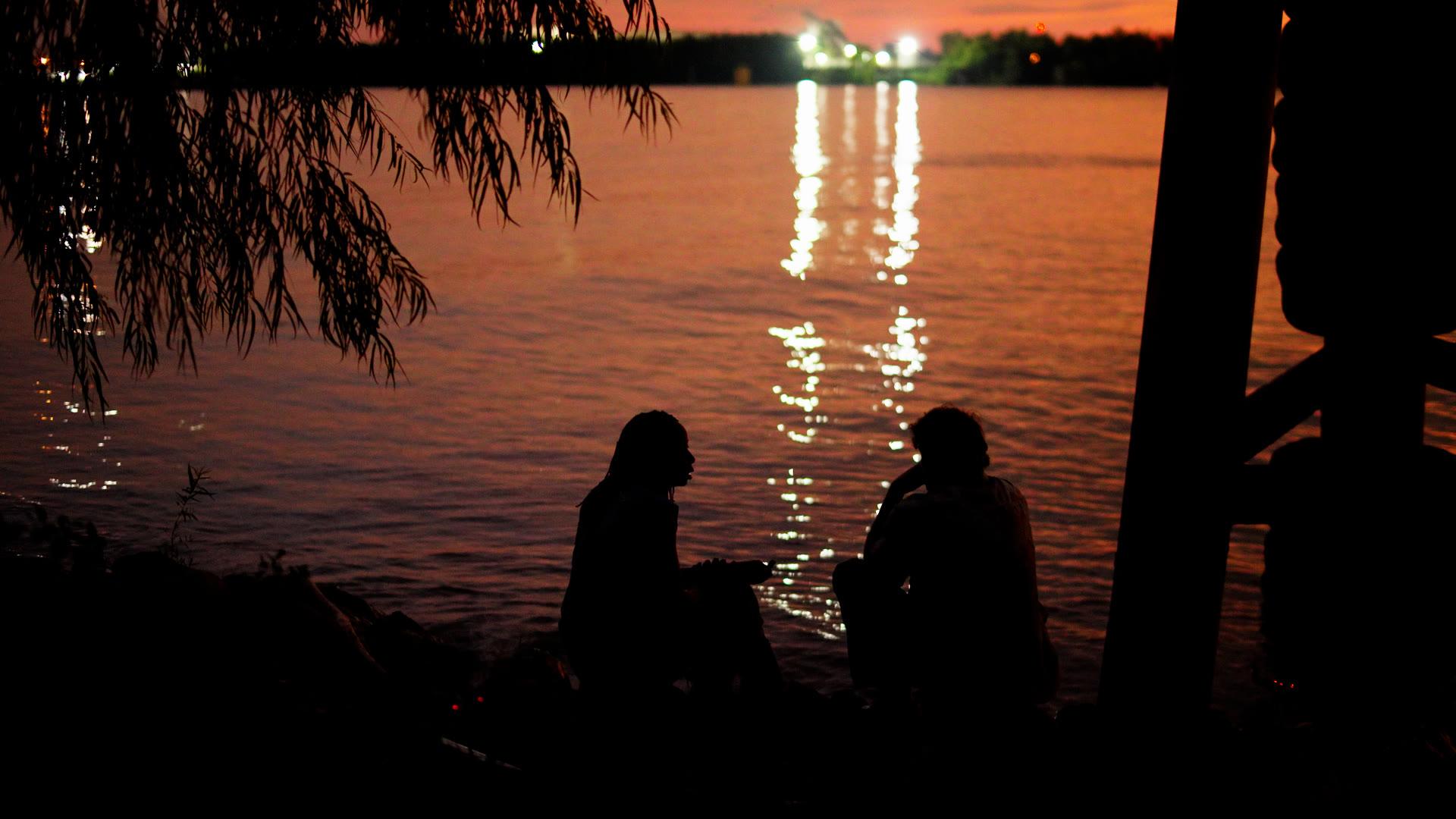 Two people in quiet conversation by the water at dusk